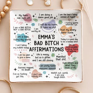 Könnte beinhalten: Ein rechteckiges Keramik-Schmucktablett mit goldenem Rand, mit dem Text "EMMA'S BAD BITCH AFFIRMATIONS" und verschiedenen Affirmationen in einer verspielten Schriftart. Das Tablett ist weiß mit farbenfrohen Akzenten.