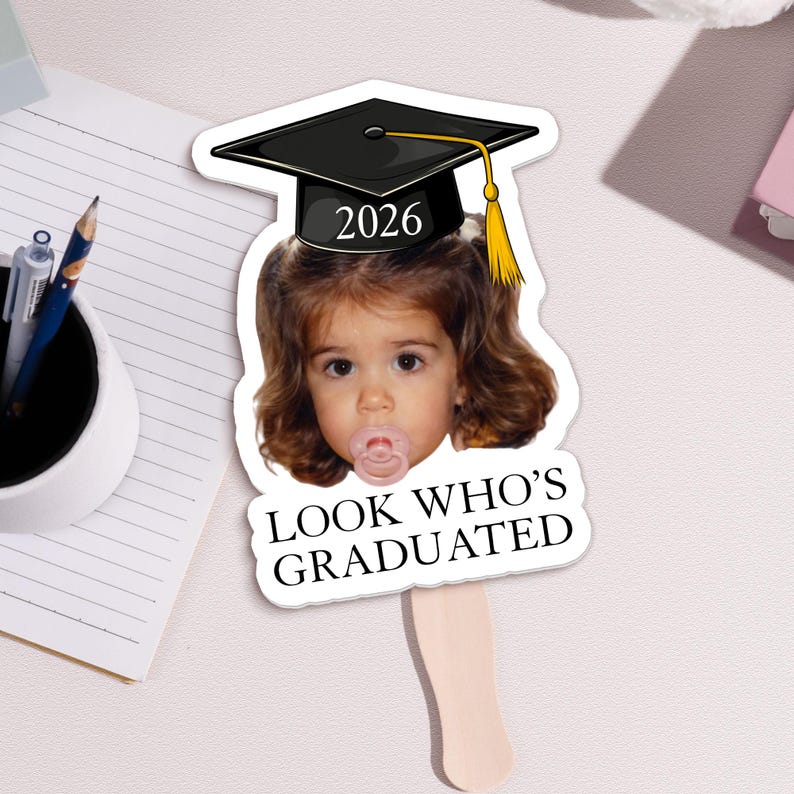 May include: A graduation-themed fan featuring a child's face wearing a black graduation cap with a gold tassel and the year 2026. The fan has the text "LOOK WHO'S GRADUATED" on the bottom.