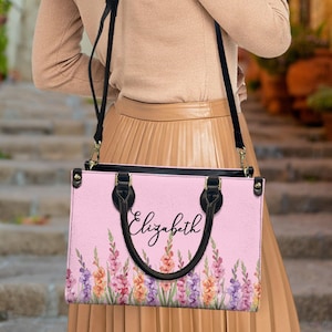 May include: A pink handbag with a floral pattern and the name "Elizabeth" in cursive. The bag has black handles and a black shoulder strap. The floral design includes purple, orange, and pink flowers.