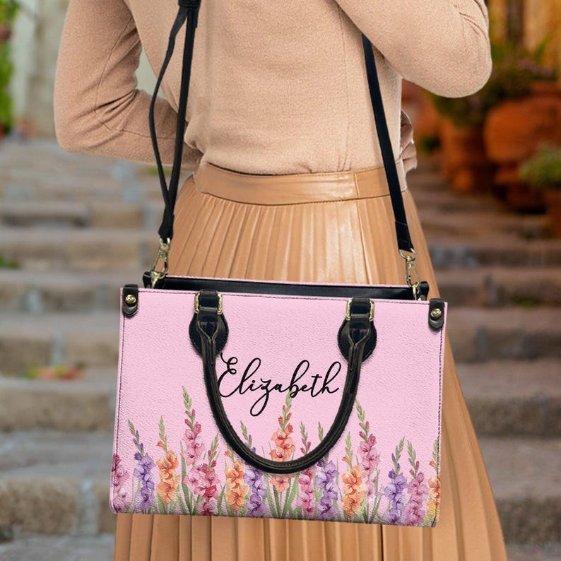 May include: A pink handbag with a floral pattern and the name "Elizabeth" in cursive. The bag has black handles and a black shoulder strap. The floral design includes purple, orange, and pink flowers.