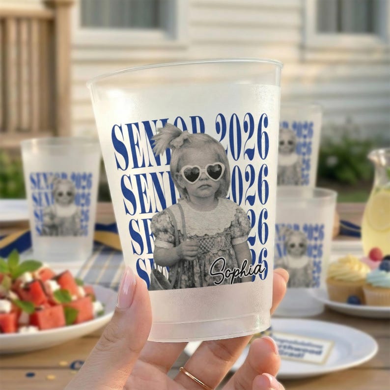 Op de afbeelding: Transparante plastic bekers met een blauw-wit ontwerp. De bekers hebben de tekst "SENIOR 2026" en een zwart-wit afbeelding van een jong meisje met een hartvormige zonnebril. De naam "Sophia" staat ook op de beker.