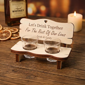 May include: A wooden shot glass holder shaped like a bench, with the text "Let's Drink Together For The Rest Of Our Lives Noah & Emily". The holder has two clear shot glasses and is on a wooden surface.