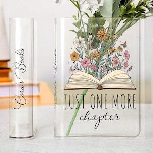 May include: Clear glass book vase with the text "Just One More Chapter" and a floral design. A smaller clear glass book vase is to the left with the text "Cora's Books". Flowers are inside the larger vase.