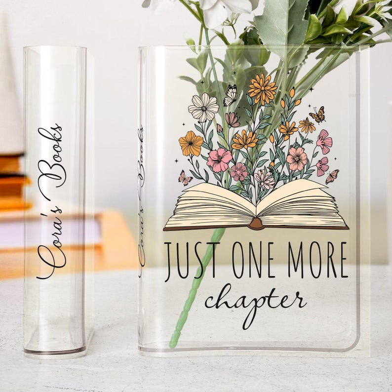 May include: Clear glass book vase with the text "Just One More Chapter" and a floral design. A smaller clear glass book vase is to the left with the text "Cora's Books". Flowers are inside the larger vase.