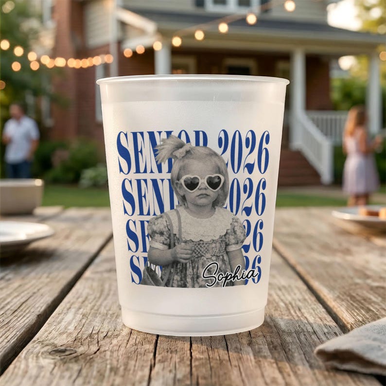 Op de afbeelding: Witte plastic beker met een blauw "SENIOR 2026" ontwerp. Het ontwerp bevat een zwart-wit foto van een kind met een hartvormige zonnebril. De naam "Sophia" staat onder de foto.