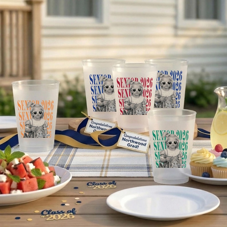 Op de afbeelding: Matte plastic bekers met de tekst "SENIOR 2026" en een foto van een kind met een zonnebril. De bekers staan op een houten tafel met eten en decoraties.