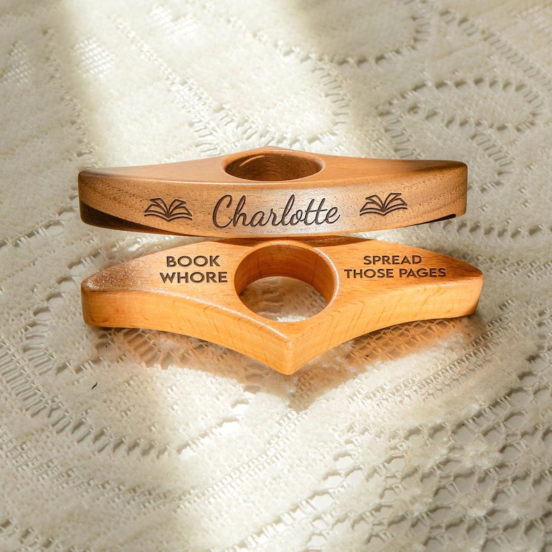 May include: Two wooden book holders. The top one is engraved with the name "Charlotte" and book illustrations. The bottom one says "BOOK WHORE" and "SPREAD THOSE PAGES". Both have a hole for the finger.