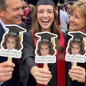 May include: A group of people at a graduation ceremony holding signs with a child's photo wearing a graduation cap. The signs read "2026" and "LOOK WHO'S GRADUATED". The graduate is wearing a cap and gown.