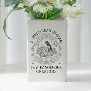 May include: A rectangular white ceramic vase with the text "A WELL-READ WOMAN IS A DANGEROUS CREATURE" and a black illustration of a woman reading. The vase is filled with white and green flowers.