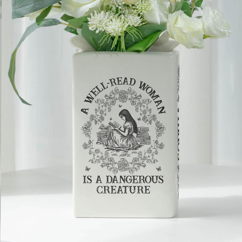 May include: A rectangular white ceramic vase with the text "A WELL-READ WOMAN IS A DANGEROUS CREATURE" and a black illustration of a woman reading. The vase is filled with white and green flowers.