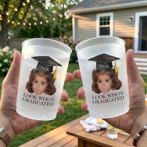 Pu&ograve; includere: Due bicchieri di plastica trasparente con un design a tema laurea. Ogni bicchiere presenta una foto di un bambino con un cappello di laurea con l'anno 2026 e il testo "LOOK WHO'S GRADUATED". I bicchieri sono tenuti davanti a uno sfondo sfocato all'aperto.