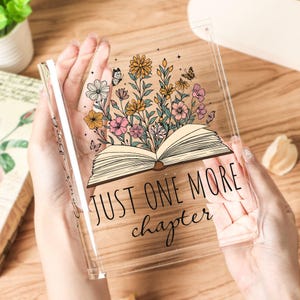 May include: Clear acrylic book-shaped decor with a floral design and the text "JUST ONE MORE chapter." The design features an open book with colorful flowers and butterflies. The acrylic piece is held in hands.