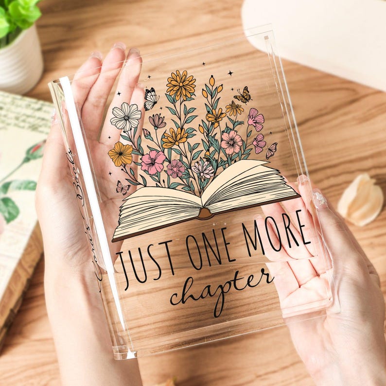 May include: Clear acrylic book-shaped decor with a floral design and the text "JUST ONE MORE chapter." The design features an open book with colorful flowers and butterflies. The acrylic piece is held in hands.