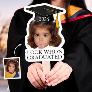May include: A graduation photo prop featuring a child's face with a pacifier, wearing a graduation cap with the year 2026. The text "LOOK WHO'S GRADUATED" is below the image. A smaller photo of the child is in the lower left corner.