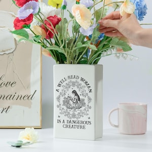 May include: A white rectangular vase with the text "A WELL-READ WOMAN IS A DANGEROUS CREATURE" and a floral illustration. The vase holds a bouquet of colorful artificial flowers. A pink and white marble mug is to the right.