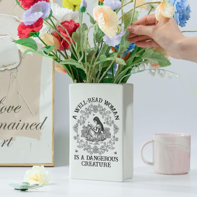 May include: A white rectangular vase with the text "A WELL-READ WOMAN IS A DANGEROUS CREATURE" and a floral illustration. The vase holds a bouquet of colorful artificial flowers. A pink and white marble mug is to the right.