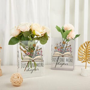 May include: Two clear rectangular vases with floral arrangements. Each vase has an open book graphic with the text "JUST ONE MORE chapter". One vase holds cream-colored roses, the other holds pink flowers. The vases are decorated with colorful flowers.