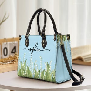 May include: Pale blue handbag with black handles and a detachable shoulder strap. The bag features a floral design with white flowers and green leaves, and the name "Sophia" in cursive. Gold-coloured hardware accents the bag's design.