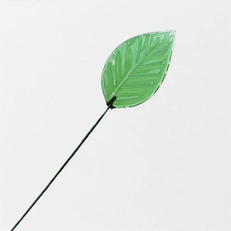 May include: A single, vibrant green leaf on a dark wire stem. The leaf has detailed veining and a glossy finish, suggesting it's made of glass or a similar material. The stem is thin and dark, contrasting with the bright leaf.