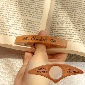 May include: A wooden book page holder with the name "Charlotte" engraved on it. The holder has a finger hole and the words "Book Whore" and "Spread Those Pages" are engraved on the sides. The holder is resting on an open book.