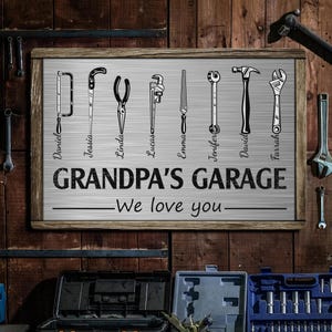 May include: A framed sign with a wooden border and a metallic background. The sign features illustrations of various tools, including a saw, pliers, and wrenches. The text reads "GRANDPA'S GARAGE - We love you" with names below the tools.