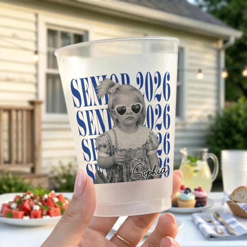 Op de afbeelding: Een doorzichtige plastic beker met een zwart-wit afbeelding van een kind met een hartvormige zonnebril. De beker heeft de blauwe tekst "SENIOR 2026" en de naam "Sophia".