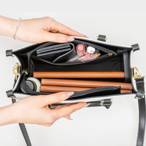 May include: Open white and black handbag with a top handle and a black shoulder strap. The interior is black and contains a wallet, cosmetics, and brown cylindrical items. Gold-tone hardware is visible.