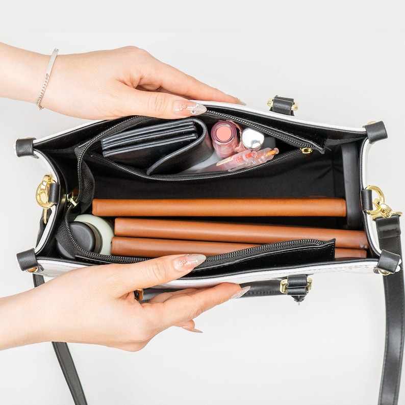 May include: Open white and black handbag with a top handle and a black shoulder strap. The interior is black and contains a wallet, cosmetics, and brown cylindrical items. Gold-tone hardware is visible.