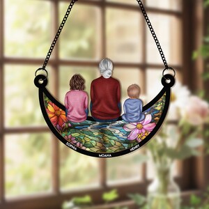 May include: A decorative family-themed hanging ornament. The ornament features a black crescent shape with a chain for hanging. The design depicts a family of three, with the names Sophia, Moana, and Henry. The background is a colorful floral and water scene.