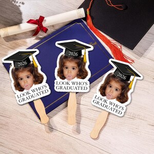 May include: Three graduation-themed photo props featuring a child wearing a graduation cap with the year 2026. Each prop has the text "LOOK WHO'S GRADUATED". A diploma and graduation cap with tassel are also visible.