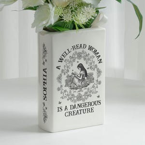 May include: A white rectangular vase with the text "A WELL-READ WOMAN IS A DANGEROUS CREATURE" and the name "SOPHIA" printed on the side. The vase features a black illustration of a woman reading and is filled with white flowers.