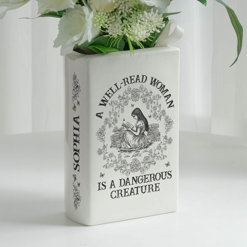 May include: A white rectangular vase with the text "A WELL-READ WOMAN IS A DANGEROUS CREATURE" and the name "SOPHIA" printed on the side. The vase features a black illustration of a woman reading and is filled with white flowers.