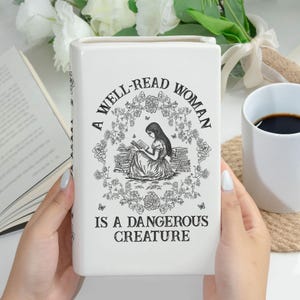 May include: White ceramic book-shaped vase with the text "A WELL-READ WOMAN IS A DANGEROUS CREATURE" and a black and white illustration of a woman reading. The vase is rectangular and has a glossy finish.