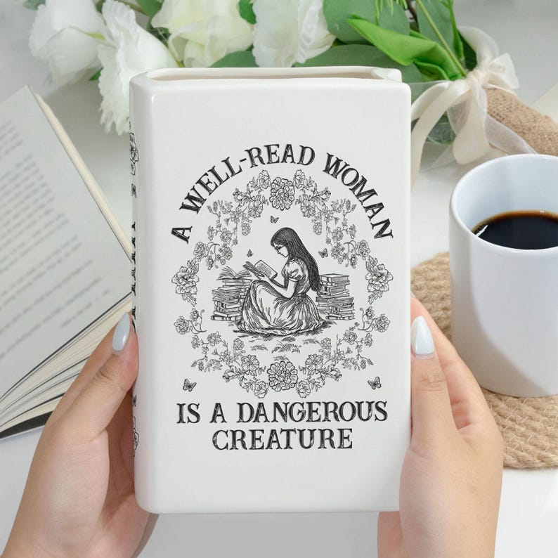 May include: White ceramic book-shaped vase with the text "A WELL-READ WOMAN IS A DANGEROUS CREATURE" and a black and white illustration of a woman reading. The vase is rectangular and has a glossy finish.