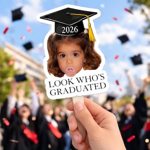 May include: A graduation-themed photo prop featuring a child's face wearing a graduation cap with the year 2026. The prop has the text "LOOK WHO'S GRADUATED" and is held in front of a blurred background of graduates.