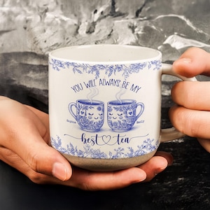 May include: A white ceramic mug with a blue floral design and the text "YOU WILL ALWAYS BE MY best tea". The mug features two cartoon teacups with smiling faces and the names "Sophia" and "Emily".