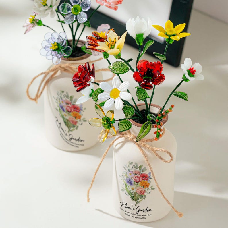 May include: Two cream-colored vases with colorful glass flowers. Each vase has a floral design and the text "Mom's Garden". The vases are tied with twine. The flowers are red, yellow, white, and clear.
