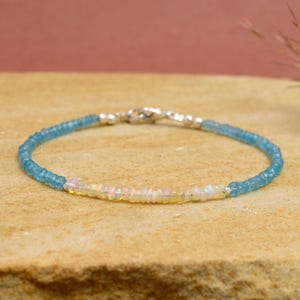 Op de afbeelding: Een verfijnde armband met een reeks kleine, gefacetteerde blauwe kralen en een middengedeelte van iriserende, melkwitte opaalkralen. Het sieraad is afgewerkt met een zilverkleurige karabijnsluiting en kleine zilveren tussenkralen.