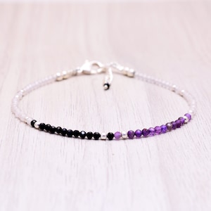 Könnte beinhalten: Ein zartes Armband mit einem Farbverlauf aus weißen, schwarzen und violetten Perlen. Das Armband besteht aus kleinen, runden Perlen und hat einen silbernen Verschluss.