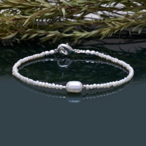 May include: A delicate white pearl bracelet with a single larger pearl accent. The bracelet is made of small white beads and has a silver clasp.