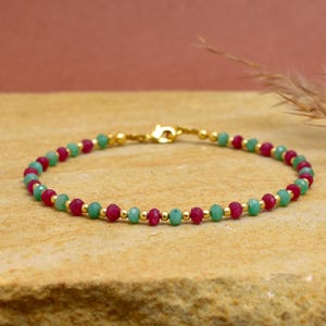 Op de afbeelding: Een delicate armband met afwisselend rode en groene edelsteen kralen, afgewisseld met kleine goudkleurige kralen. De armband is vastgezet met een goudkleurige sluiting. De sieraden worden gepresenteerd op een neutraal oppervlak.