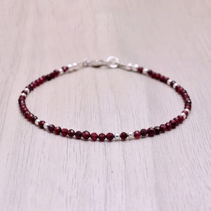 May include: A delicate bracelet made with small, faceted garnet beads and silver beads. The bracelet is on a wooden surface.