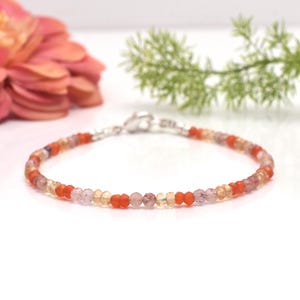 Op de afbeelding: Een delicate armband gemaakt met kleine, ronde kralen in tinten oranje, roze en helder. De kralen zijn geregen aan een zilveren ketting met een karabijn sluiting.