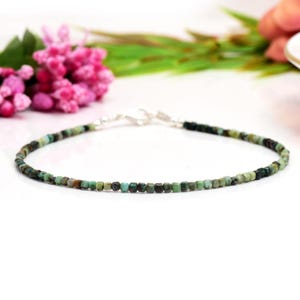 Può includere: Un braccialetto delicato con piccole perline quadrate nei toni del verde, marrone e nero. Le perline sono infilate con una chiusura color argento. Il braccialetto è presentato su una superficie bianca, con fiori rosa e fogliame verde sullo sfondo.