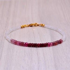 May include: A delicate bracelet with a gradient of pink and red beads, interspersed with white beads. The bracelet has a gold clasp.