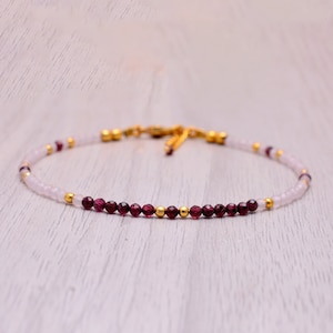 May include: A delicate gold chain bracelet with alternating white and burgundy beads. The bracelet is made with small, round beads and has a simple, elegant design.