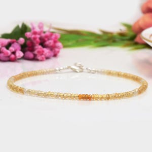 Op de afbeelding: Een delicate armband van gouden en oranje edelstenen met een zilveren sluiting. De armband is gemaakt van kleine, ronde kralen en is perfect om te combineren met andere armbanden.