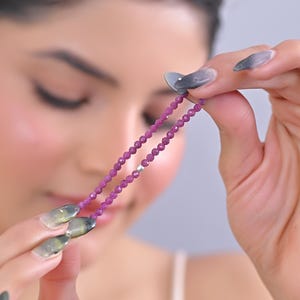 May include: Two strands of small, faceted, deep pink gemstones are held up against a soft background. The beads are strung together, creating a delicate necklace or bracelet. The gemstones have a subtle sparkle.