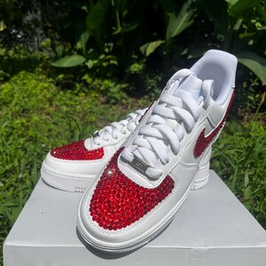 May include: White sneakers with red jewel embellishments on the toe and Nike swoosh. The shoes have white laces and a white sole. The sneakers are sitting on a white box, with a green background.