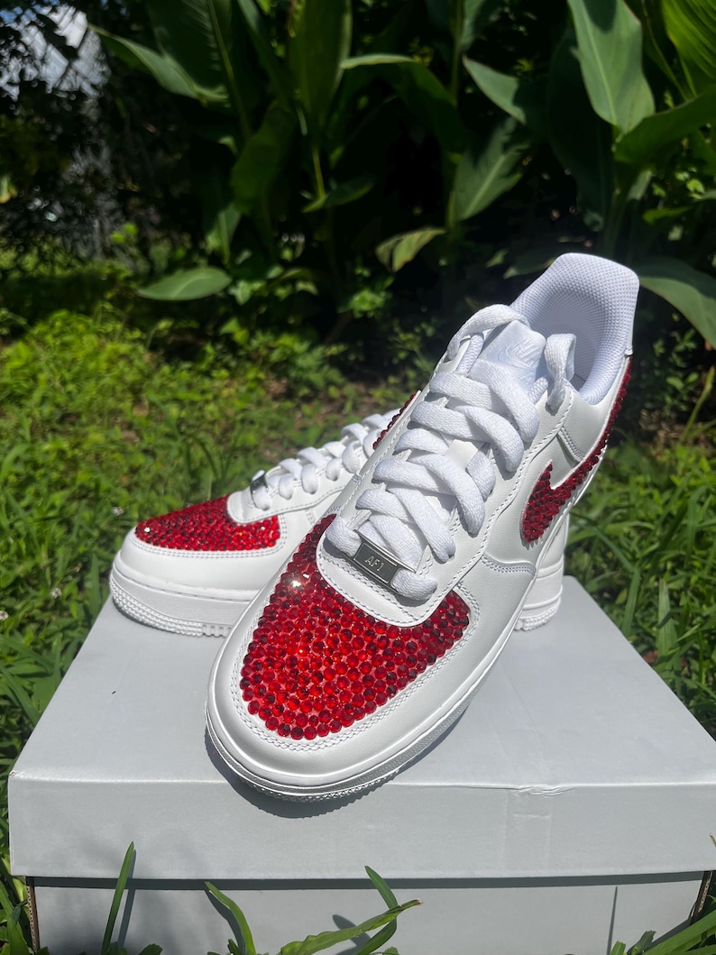 May include: White sneakers with red jewel embellishments on the toe and Nike swoosh. The shoes have white laces and a white sole. The sneakers are sitting on a white box, with a green background.